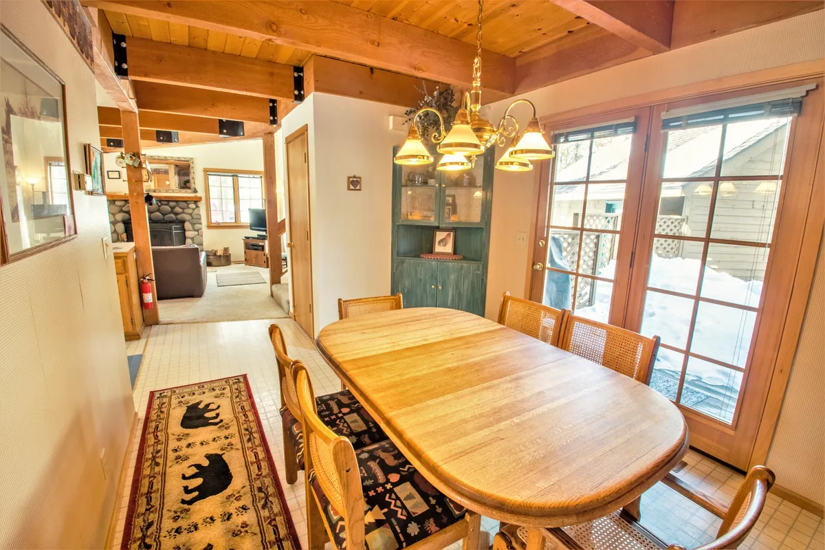 A warm and inviting dining area with a wooden table set beneath a striking chandelier, surrounded by natural wood beams and large windows that let in plenty of daylight. The space is framed by the textures of wood and soft light, evoking a sense of cozy intimacy ideal for shared meals or quiet mornings with coffee.