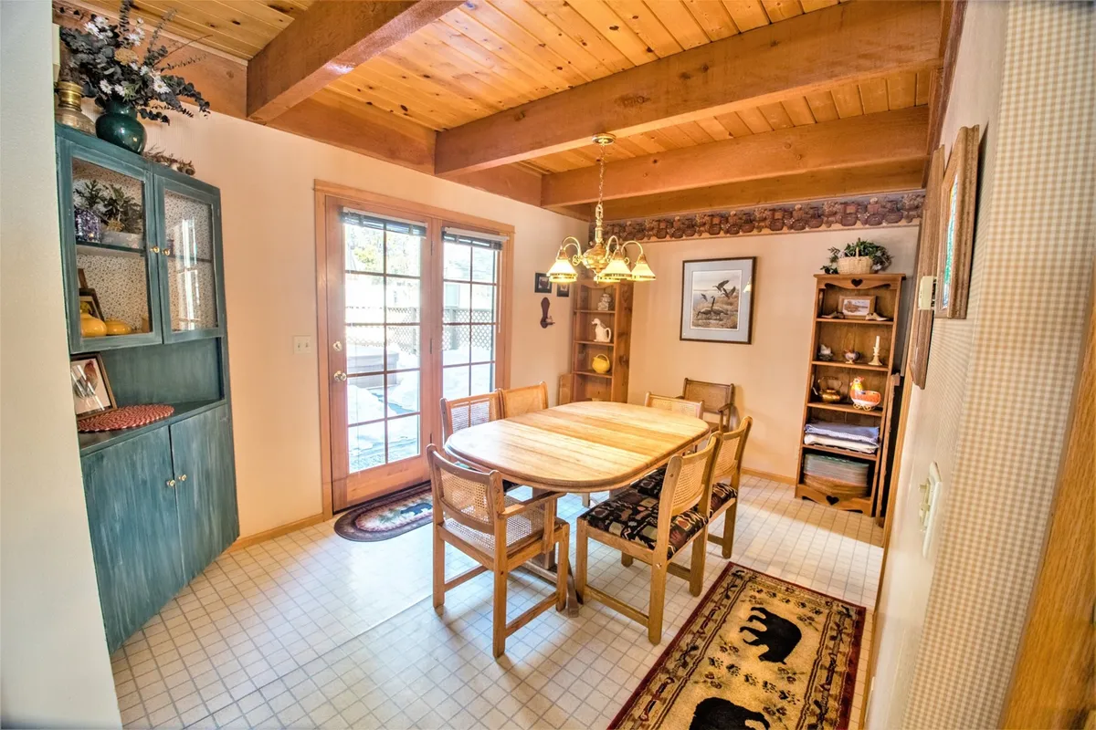 The image focuses on a warm dining area set beneath a rustic chandelier, with wooden beams overhead and a sliding glass door letting in natural light. The table is the focal point, ready for family meals, celebrations, or casual brunches. A bear-motif rug adds a touch of mountain charm.