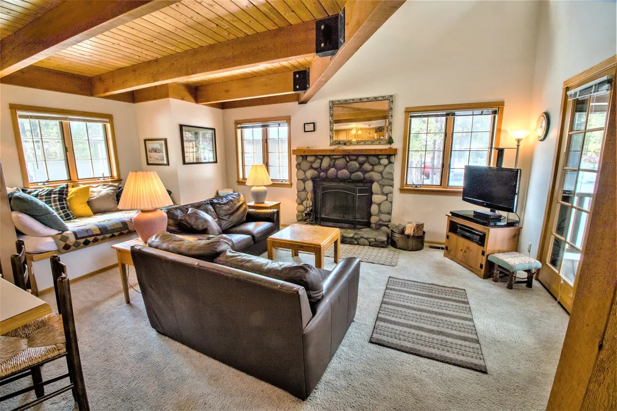 The image showcases a warm and inviting living room with a rustic stone fireplace as the centerpiece, comfortable seating arranged for conversation and relaxation, large windows letting in forest views and natural light, and a decorative lamp adding to the cozy ambiance. The natural wood details on the ceiling and walls contribute to the cabin-in-the-woods feel, while the overall setting is perfect for unwinding, reading, or socializing with friends and family.