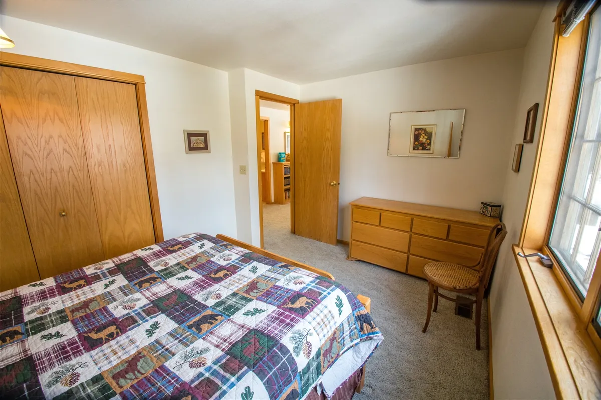 A warm, inviting bedroom featuring a queen bed adorned with a patterned quilt, a wooden dresser, an accent chair beside the window, and a doorway leading to another room. Morning sunlight streams in, enhancing the tranquil ambiance and highlighting the cozy reading nook.