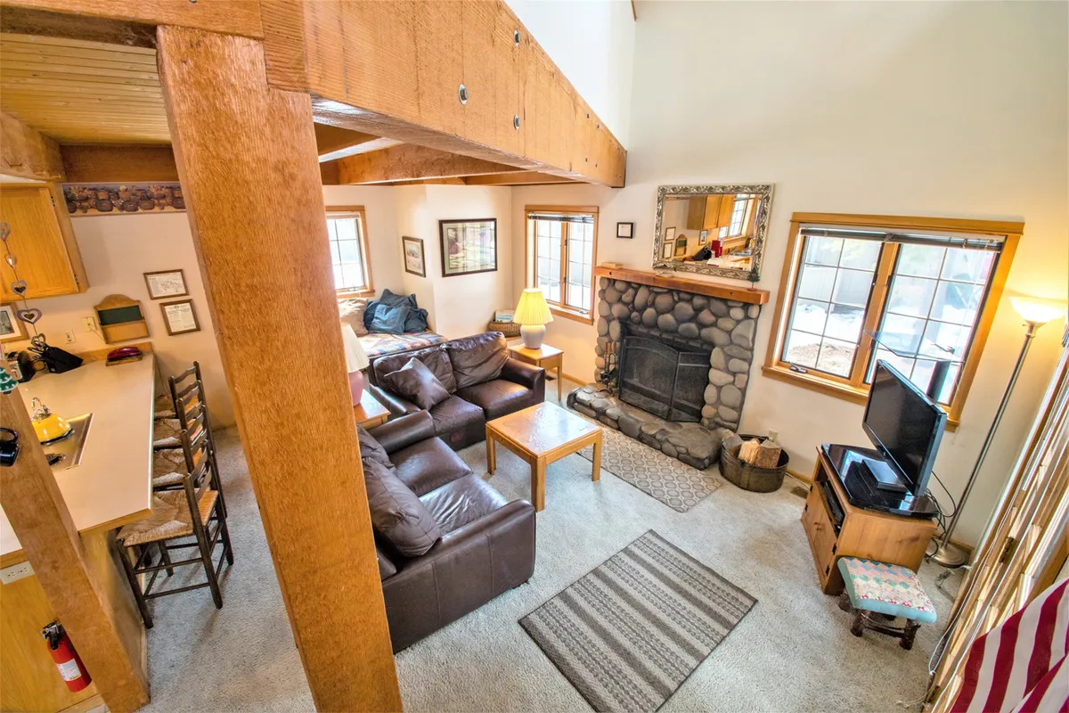 This image captures a spacious living room with a striking stone fireplace as the focal point, flanked by plush leather sofas and accented by exposed wooden beams. Large windows bathe the space in natural light, enhancing the room's warm, inviting feel. The arrangement of comfortable seating suggests options for socializing, relaxing, or gathering for a cozy movie night.