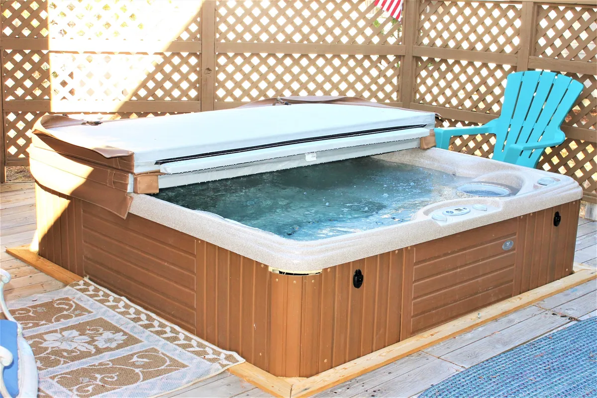 A serene deck scene featuring a bubbling hot tub surrounded by a lattice fence, blue Adirondack chair, and an outdoor rug. The space invites relaxation, with the hot tub as the focal point for unwinding under the open sky.