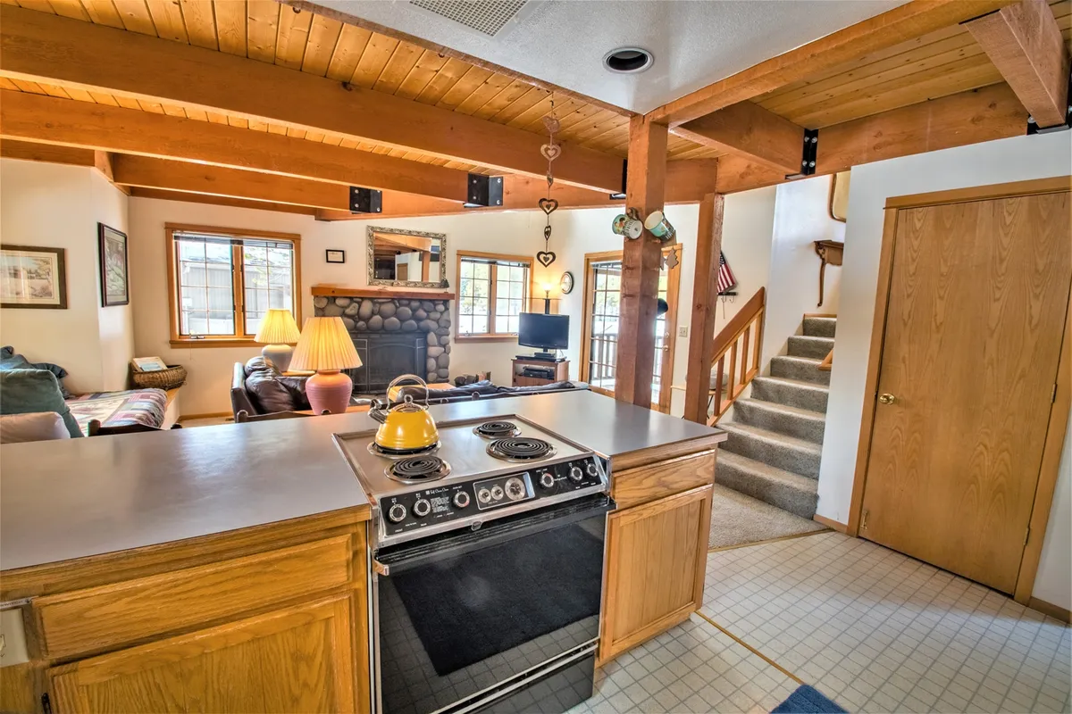 A wide shot showcasing a warm, open-concept living area where the stone fireplace, kitchen island, and comfortable seating area blend seamlessly. Wooden beams and a staircase add lodge character, while the layout highlights the open flow ideal for families and friends to gather, dine, and relax together.
