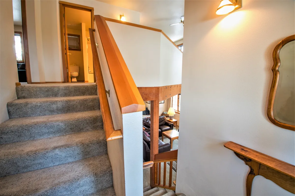 A view of the staircase with rustic wooden railings, leading upstairs, with a glimpse of a cozy seating area in the background. Morning light spills in, illuminating the inviting textures and open layout, suggesting ease of movement and social connection within the home.